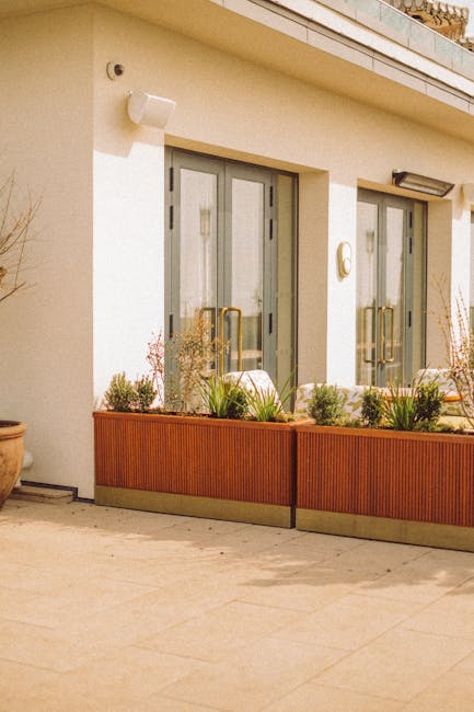 The image depicts a modern balcony garden attached to a white building, featuring a long, rectangular wooden planter box filled with various green plants, grasses, and small shrubs. Behind the planter