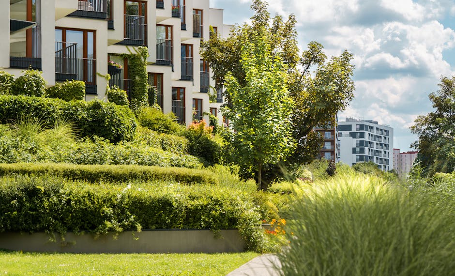 The image depicts a modern balcony garden attached to a white building, featuring a long, rectangular wooden planter box filled with various green plants, grasses, and small shrubs. Behind the planter