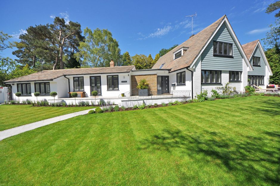 A modern, two-storey house with a white exterior and grey roofing situated in a landscaped garden. The garden features a neatly mowed green lawn with visible mowing patterns, a narrow curved concrete 